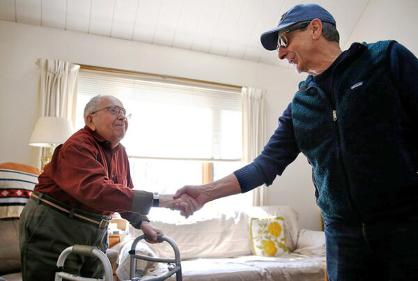 A Meals on Wheels volunteer shakes hands with an elderly man using a walker inside his living room during a home-delivered meal visit.