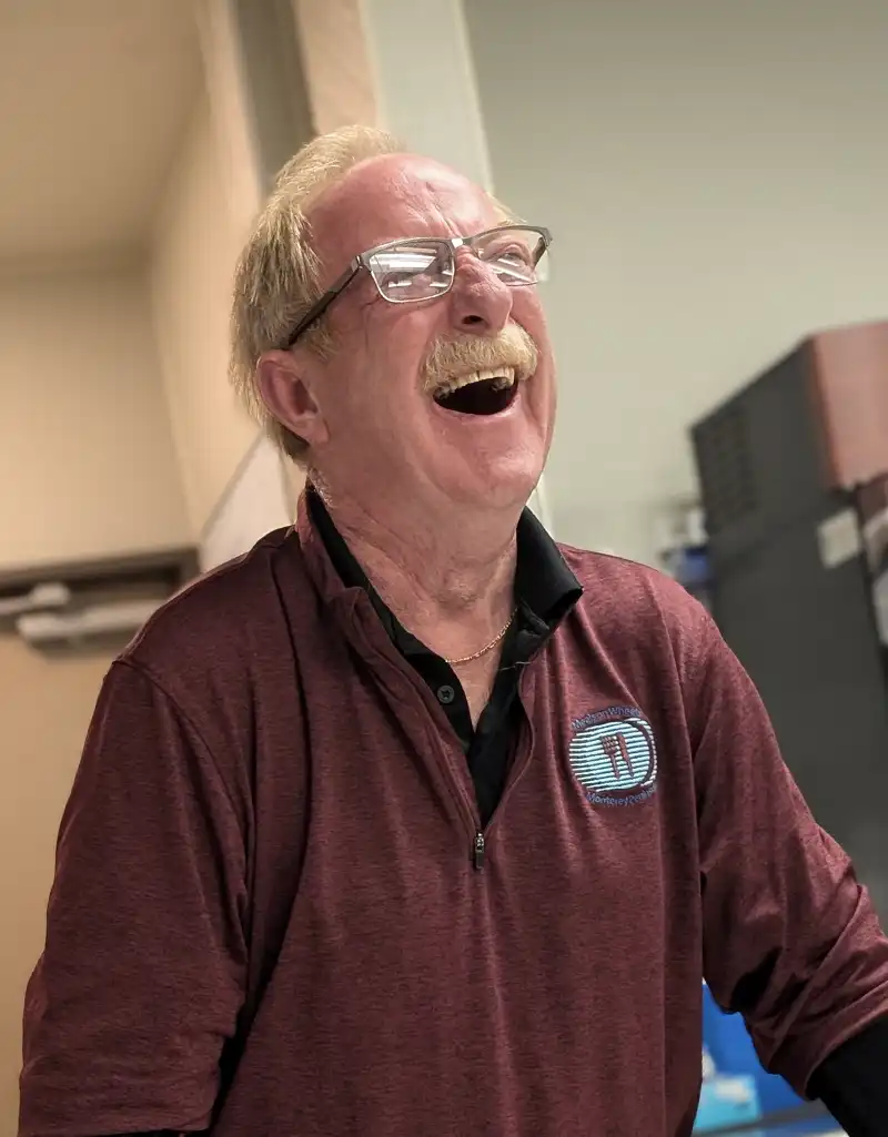 Senior Operations Director Mark Hansen laughing while wearing a Meals on Wheels of the Monterey Peninsula pullover inside the Community Center.
