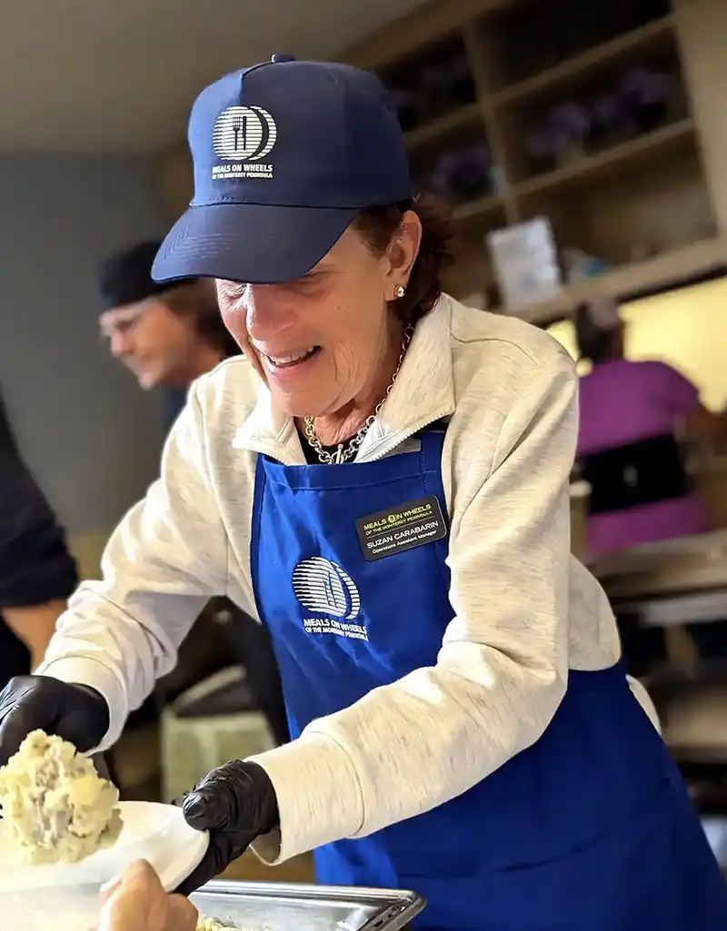Operations Manager Suzan Carabarin serving a meal to group dining guests while wearing a Meals on Wheels of the Monterey Peninsula cap and apron.