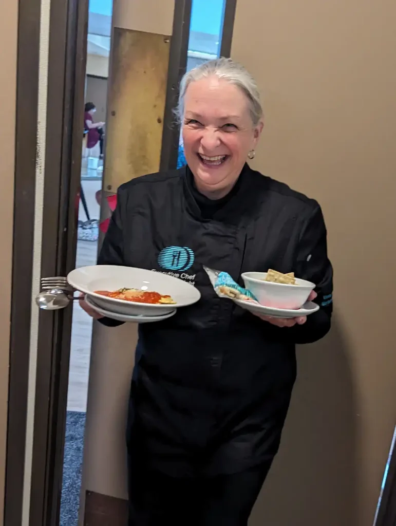 Controller Kay Smith smiling while holding plated meals, wearing a Meals on Wheels chef jacket inside the Meals on Wheels Community Center.